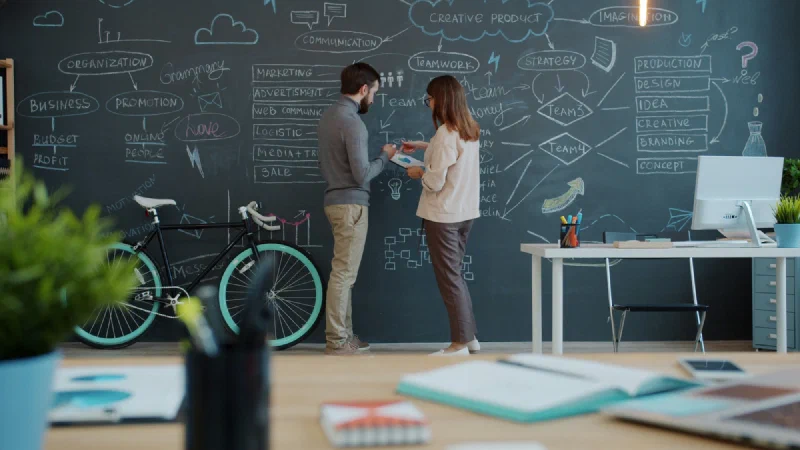 Two people planning a small business at a chalkboard wall covered in notes on budget, marketing and sales